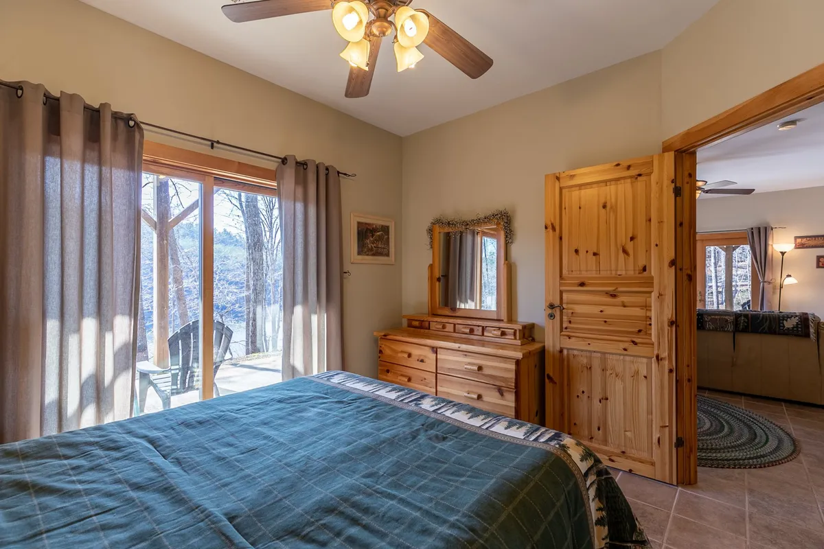 A bright bedroom with green and white bedding, a large window showcasing outdoor views, wooden furniture, and a decorative mirror, creating a warm and inviting atmosphere.