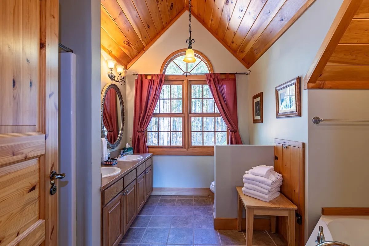 A bathroom with double vanity sinks, ample natural light from a large window, wooden ceiling and walls, a decorative mirror, and neatly folded towels, exuding tranquility and warmth.