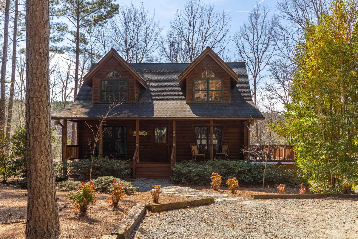 The log cabin structure is framed by large windows and surrounded by trees and a landscaped garden, creating a cozy, welcoming atmosphere. The porch hints at leisurely outdoor moments, while the environment immerses the viewer in nature.