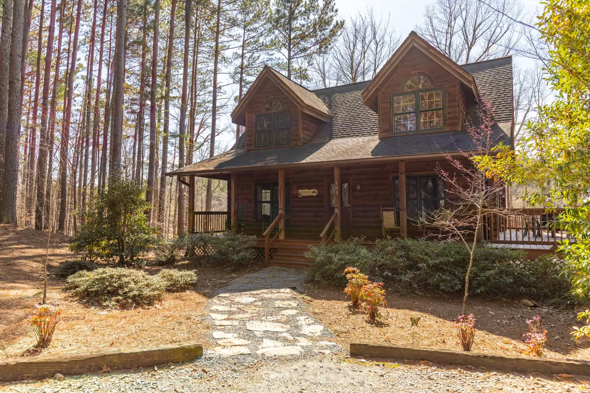 A cozy log cabin with a welcoming front porch, stone pathway, and towering trees all around. The porch is ideal for relaxing after a day on the trails, and the surrounding landscape hints at accessible walking paths and nature exploration.
