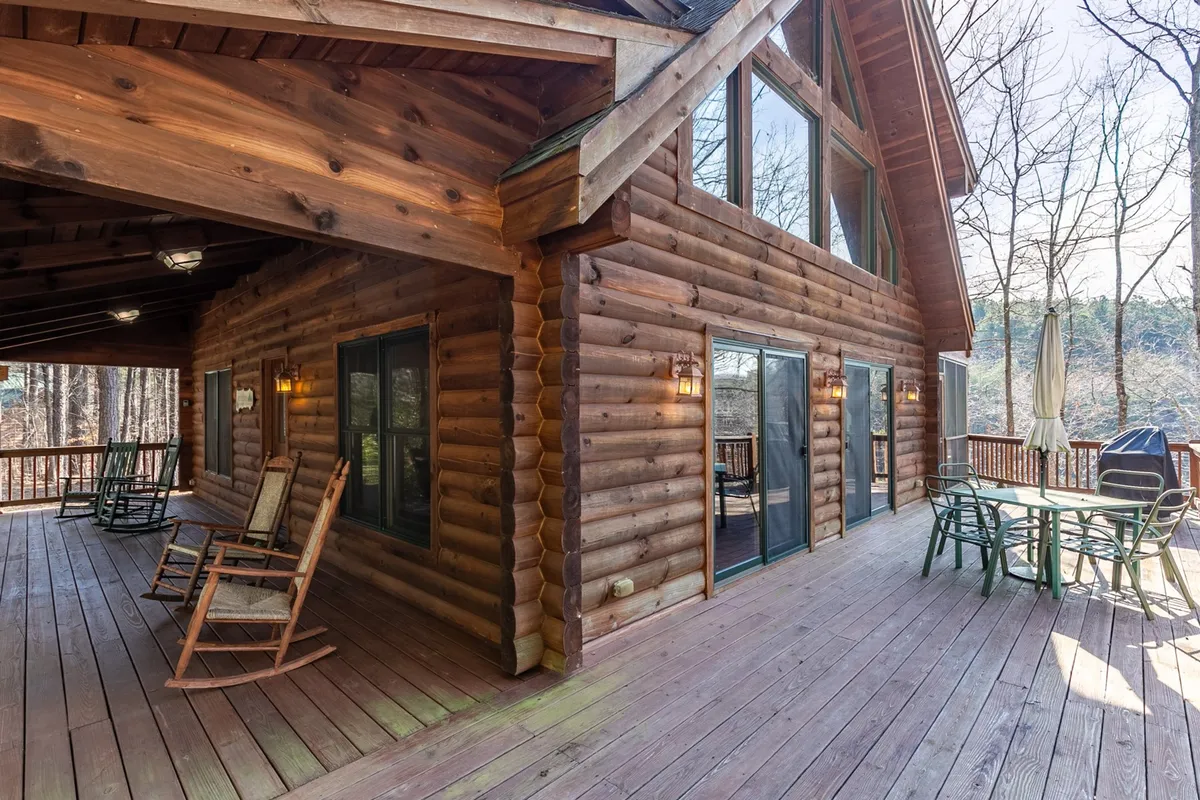 The log cabin exterior features rocking chairs and an outdoor dining set on a spacious porch. Large windows offer glimpses inside, while surrounding trees create a private, tranquil setting ideal for relaxing or socializing. The scene captures the home's seamless blend with nature and its suitability for both casual and festive gatherings.