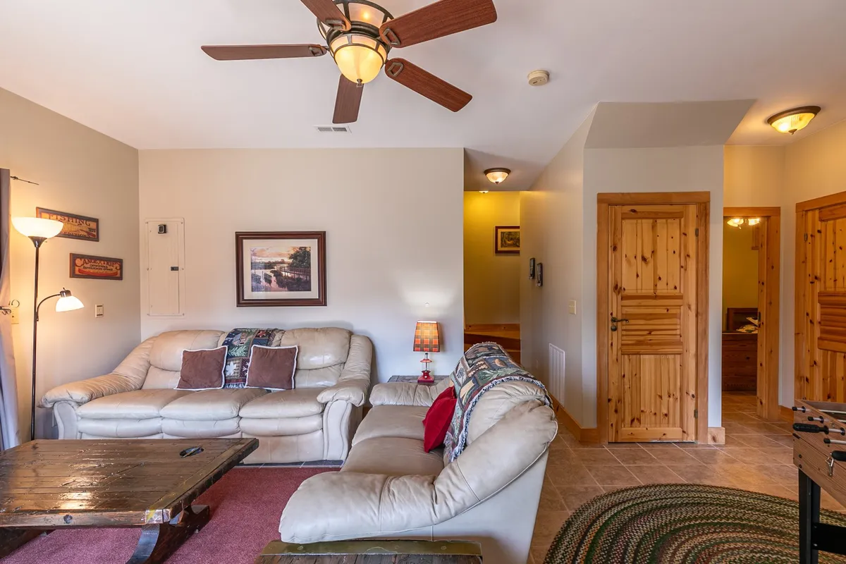 The den features plush leather sofas arranged around a wooden coffee table, with a ceiling fan and decorative lamp overhead. This inviting space is ideal for movie nights, board games, or quiet relaxation, reinforcing the comfortable, entertainment-focused atmosphere described in the section.