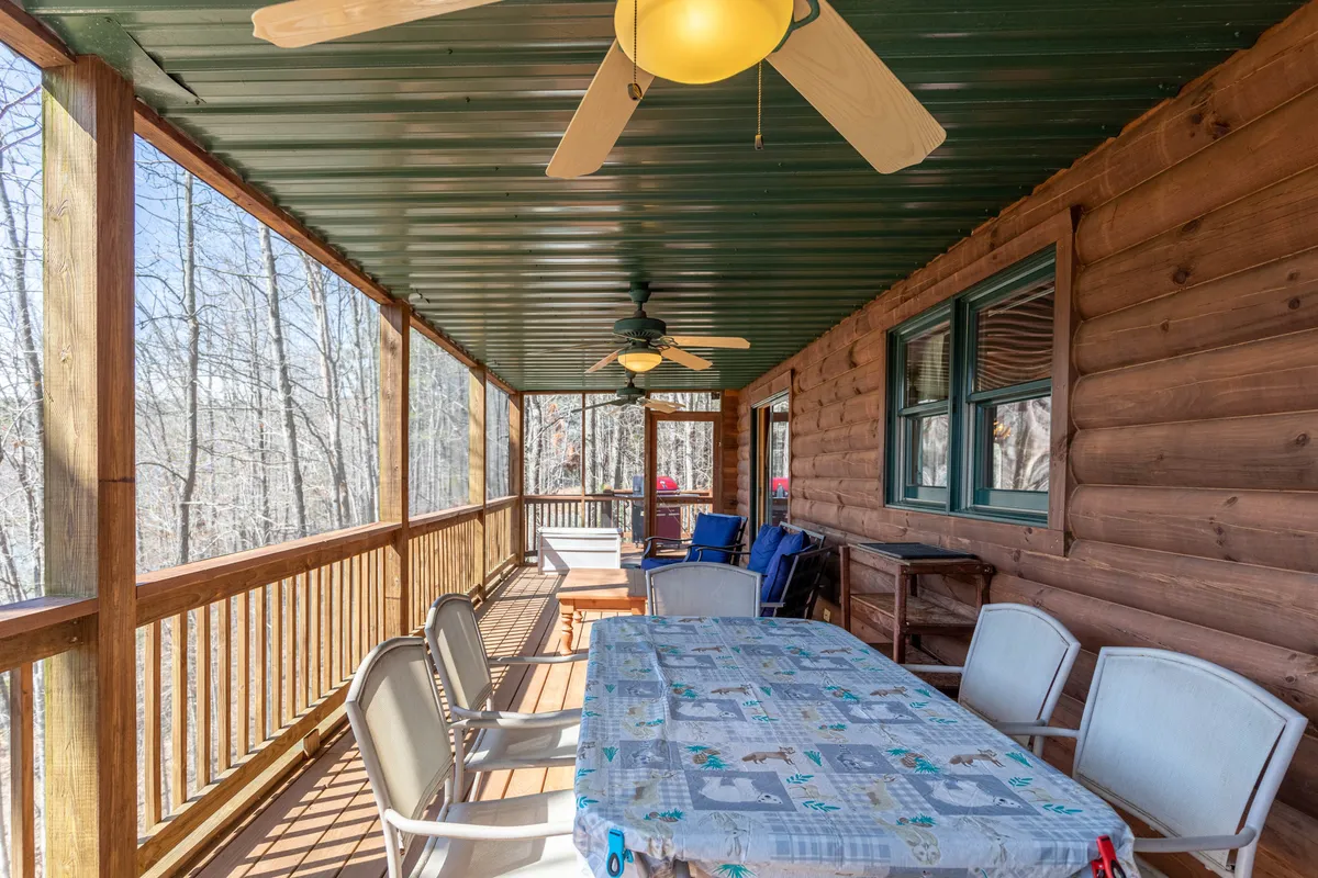 A spacious outdoor dining table sits beneath a ceiling fan on a screened porch, surrounded by leafy trees. The wooden railing frames the cozy al fresco dining area, inviting guests to gather for meals or conversation. The setup offers a comfortable transition between indoor and outdoor living, perfect for group gatherings.