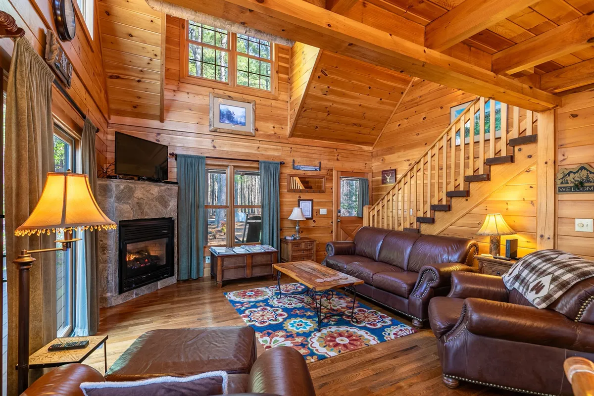 A warmly inviting interior living space featuring a sofa, fireplace, coffee table, and large windows that let in abundant natural light. The scene highlights a comfortable area ideal for relaxation or quiet moments, with the staircase hinting at the presence of an upper loft area nearby.