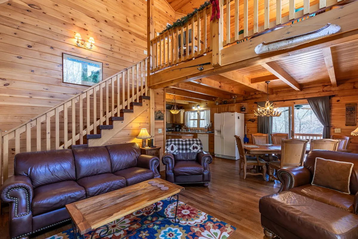 This image showcases an inviting interior with plush leather sofas arranged around a wooden dining table, a staircase, large windows flooding the space with natural light, and a statement chandelier overhead. The scene radiates warmth and provides a clear view of both the living and dining areas—spaces meant for gathering and relaxation.