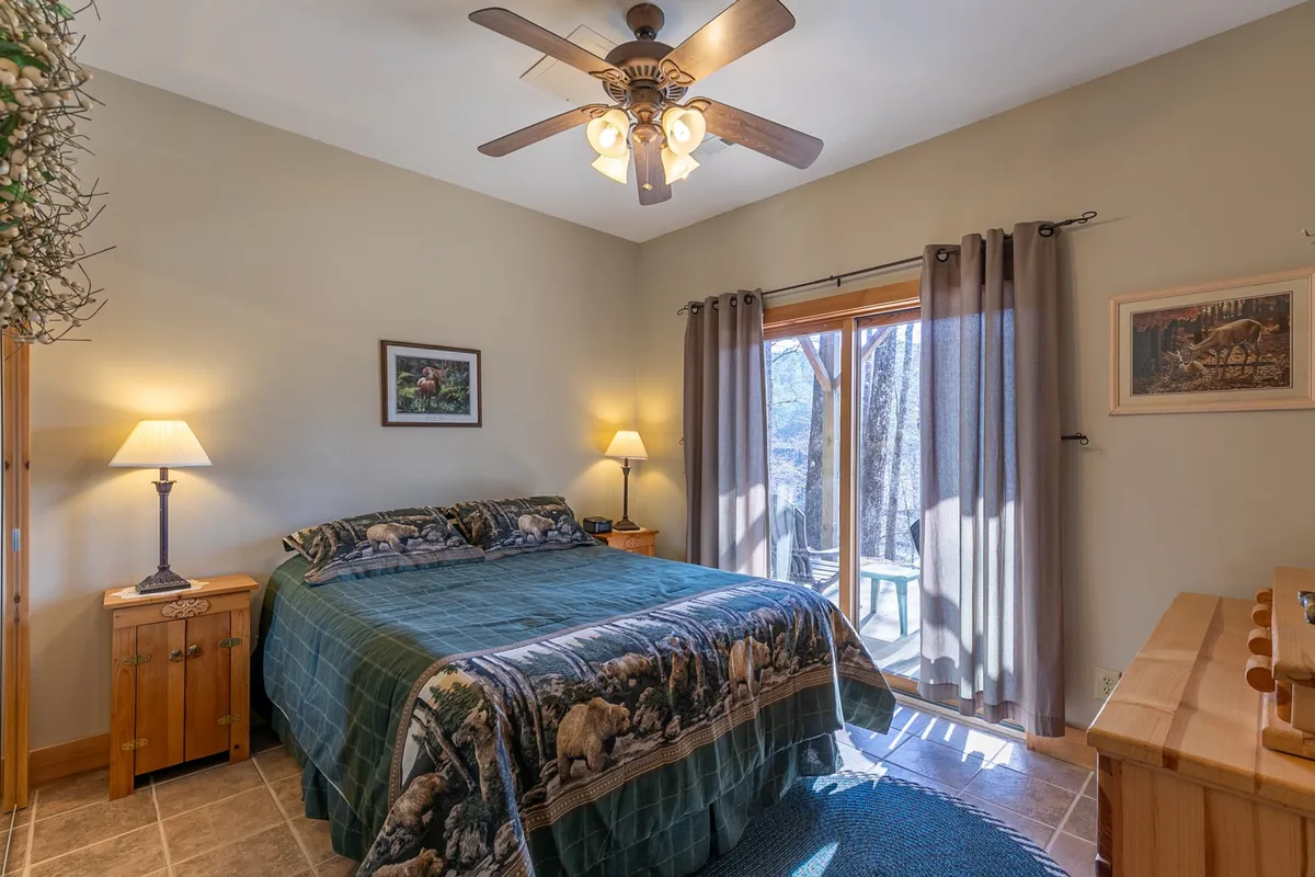 A cozy bedroom featuring a bed with animal-themed bedding, a nightstand with a lamp, and a large window that lets in natural light and offers views of the surrounding nature.