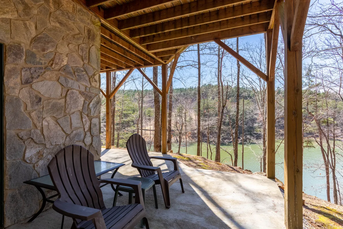 A tranquil wooden deck overlooking a serene lake, with Adirondack chairs offering a peaceful spot to relax and take in the views. The rock wall and surrounding trees enhance the sense of privacy and seclusion, encapsulating the natural beauty and quietude of the lakeside setting.